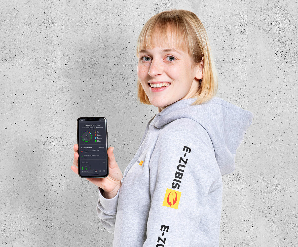 Blondes Mädchen in einem grauen Pullover mit Logo des Elektrohandwerks hält Smartphone in der Hand auf dem die Azubi-App des Elektrohandwerks geöffnet ist